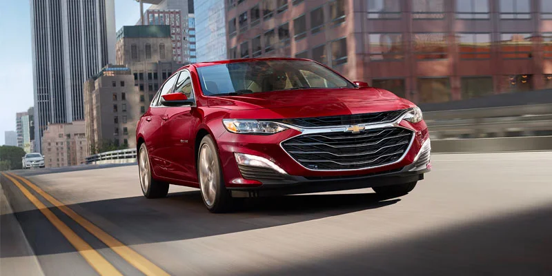 A front-facing photo of a red 2025 Chevy Malibu driving through a city near Frederic, WI