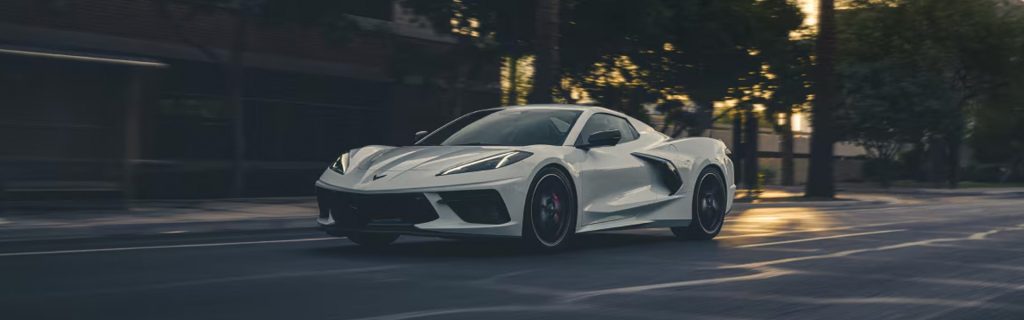 A white Chevrolet Corvette C8 driving on a street with a sunset backdrop.
