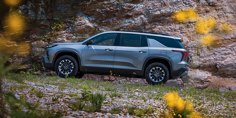 A silver Chevrolet Traverse SUV parked near a rocky cliff with some yellow flowers in the foreground.
