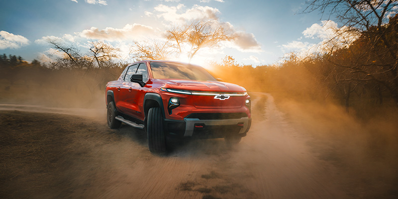 A red Chevrolet electric truck driving through a dusty trail with a sunset backdrop.
