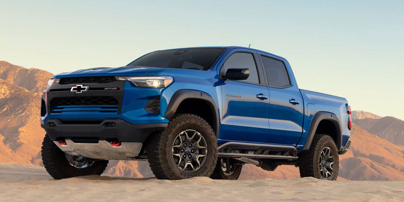 A blue Chevrolet truck parked on sandy terrain with mountains in the background.
